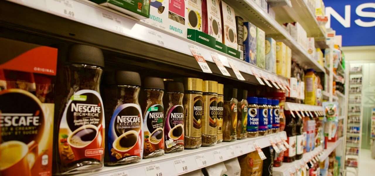Supermarktregal mit klassischen Papier-Preisschildern und Produkten wie Nescafé und Starbucks Kaffee.