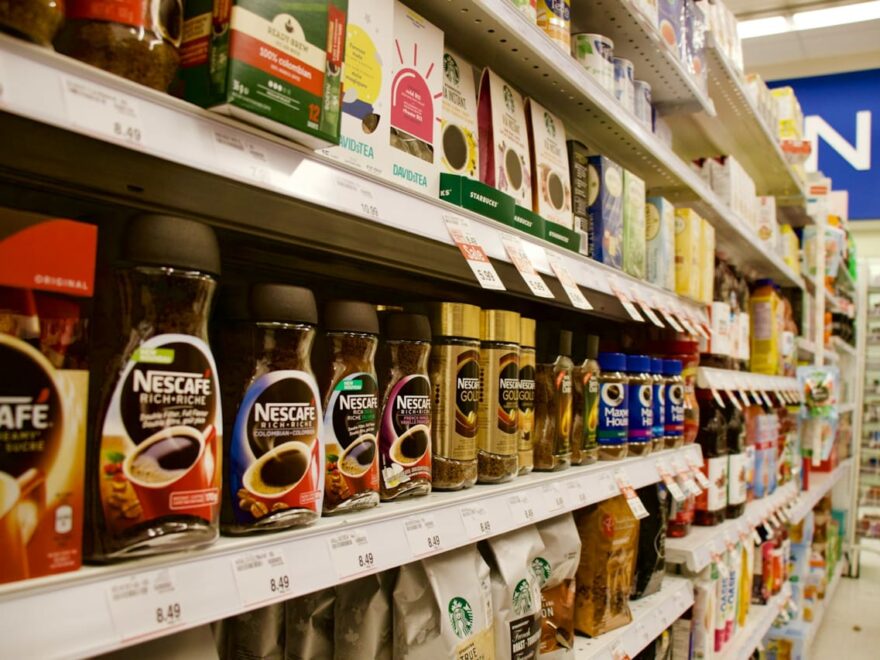 Supermarktregal mit klassischen Papier-Preisschildern und Produkten wie Nescafé und Starbucks Kaffee.