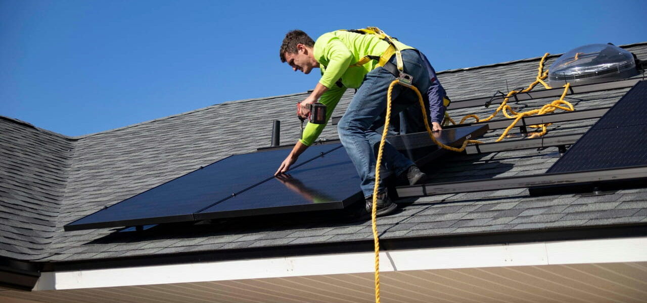 Ein Techniker installiert Solarmodule auf einem Hausdach unter klarem Himmel.